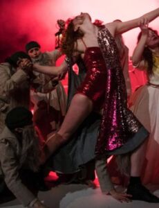 Photo du spectacle Bruegel. Une femme en robe rouge est sur scène, entourée de plusieurs personnes en gris qui lui attrapent les bras et les jambes.